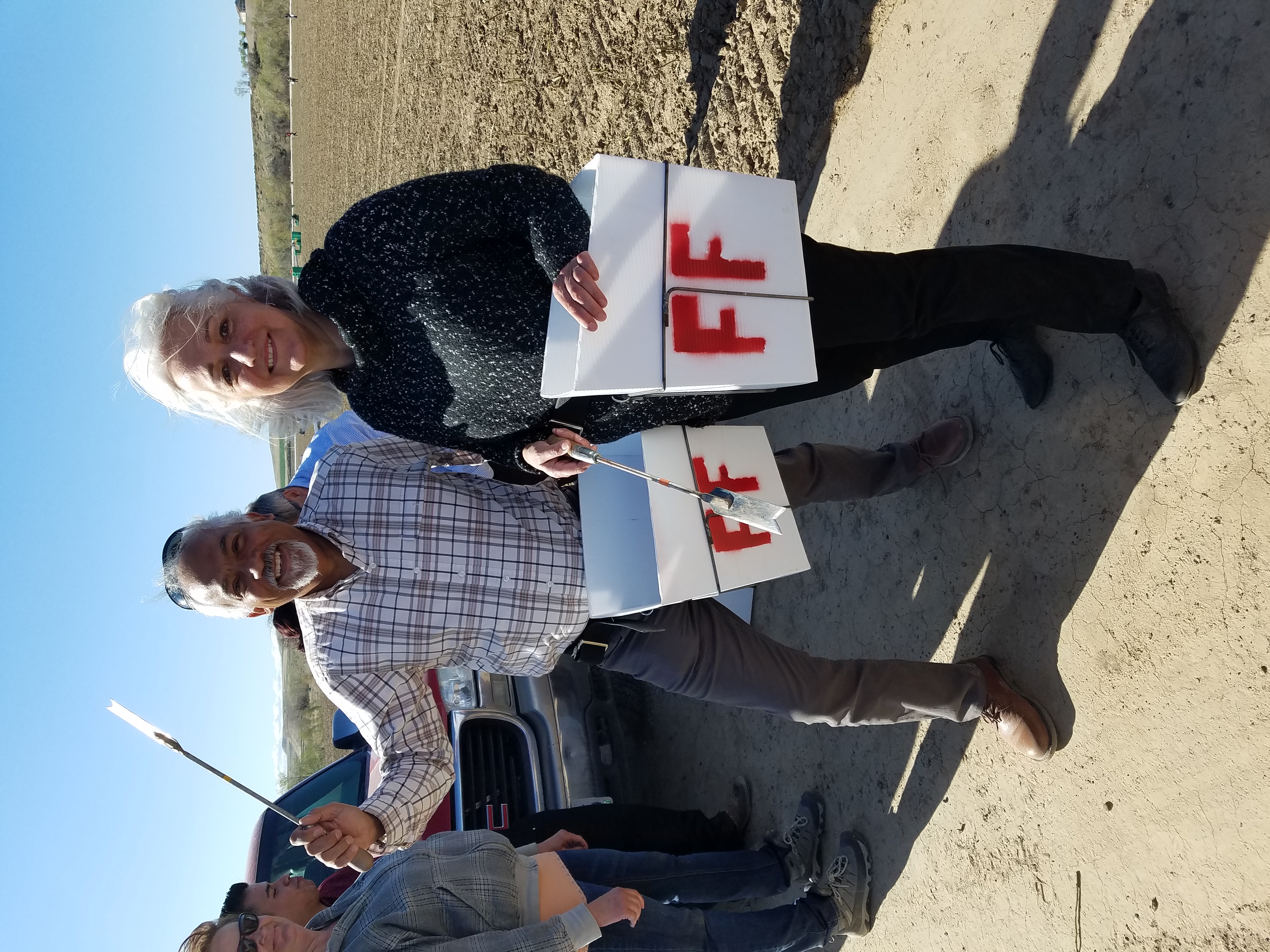 Anne Kubisch and Roque Barros stand in front of a pickup truck in an agricultural field. They each hold metal asparagus-picking tools and a box printed with the letters FF.